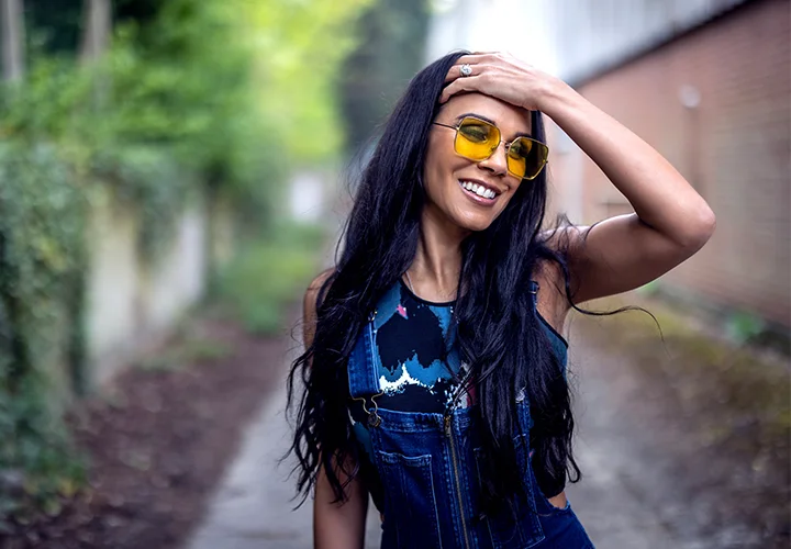 A woman wearing yellow glasses stand outside in the sun with her hands on her head smiling in blue and white shirt Adrianna Temple