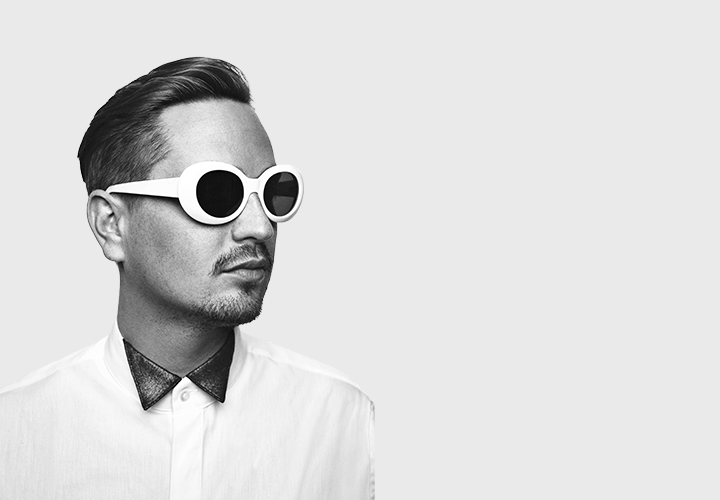 A man poses on a white background wearing white shades, black shirt and white jacket. Robin Schultz Sugar Radio