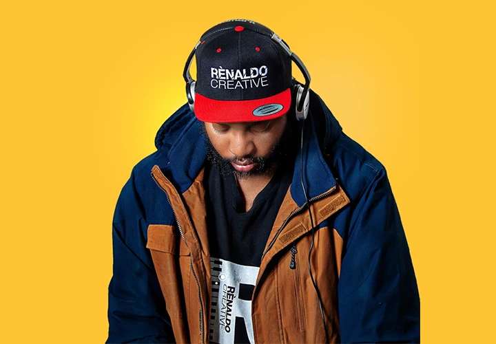 A black man looks down wearing a black and red hat with Renaldo Creative on it. Silver and black headphones. Brown and blue coat on a yellow background for Grind Hard Mix Show and Caribbean Vibes Radio Show.