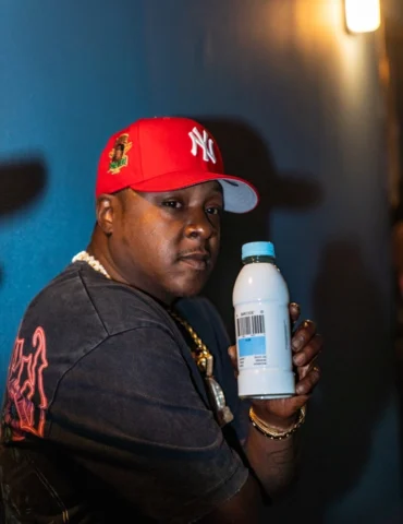 A image of Hip-Hop legend Jadakiss from Hip-Hop group Lox sittinfg in a chair wearing a D-Block chain, red New York Yankees Fited hold his energy drink. With a intenese look on his face.