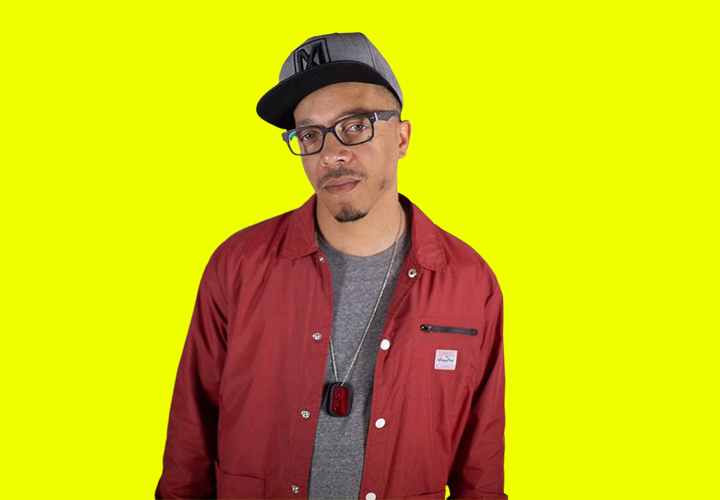 A black man Chilly Mack wears a grey hat with glasses and a button up shirt and grey shirt.