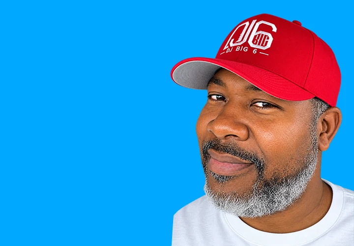 A black man with a red DJ Big 6 hat smiles in a white T-Shirt.
