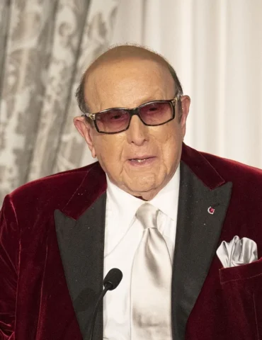 Legendary record executive Clive Davis poses in a burgany jacket, white shirt, tie purple, glasses.