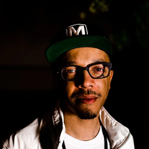 A man look at the camera wearing glasses in a Black M Hat and white shirt. 10.15.26