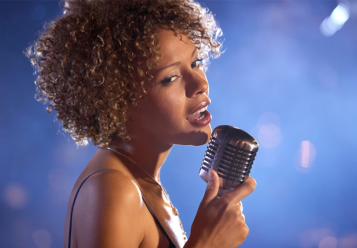A beautiful black woman with curly hair poses in a club with a mic singing on a bokeh gradient blue background. Image for Inspir3 Groove Jazz Show.